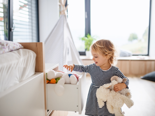 Dziewczynka wkładająca zabawki, do dodatkowej szuflady, przy łóżku dziecięcym.